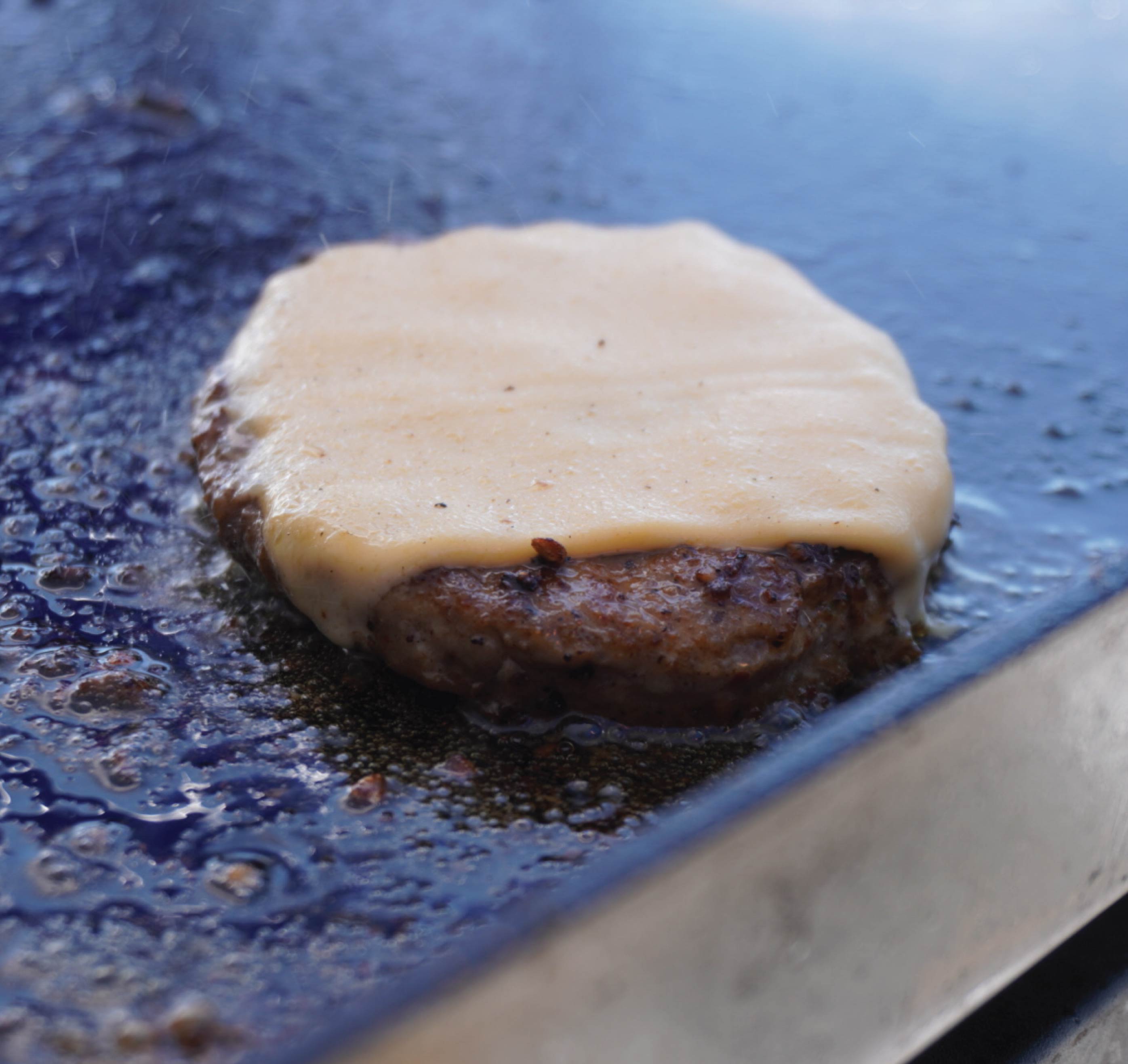 A closeup photo of a Slappy McPappy Burger patty cooking on the grill with a slice of cheese being melting on top.